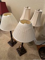 Four table lamps arranged in groups, two with multiple lampshades each shown standing on carpeted floor against a wall.