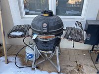 Front view of black ceramic kamado grill on metal stand with side shelves and propane tank visible.