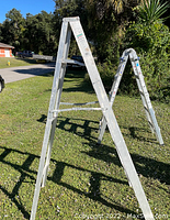 Two heavy-duty aluminum multi-position ladders standing outdoors on grass. One is in A-frame shape, the other folded.