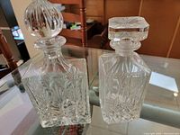 Two square etched crystal spirit decanters on glass table, showing detailed cut and etched floral and geometric patterns. One decanter has a round ribbed stopper, the other has a square stopper with starburst design.
