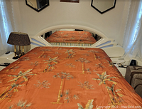 Full view of the queen size bed with large mirror headboard reflecting orange bedspread with palm pattern. Shows white lacquered wood with black inlay on headboard, side tables, and base.