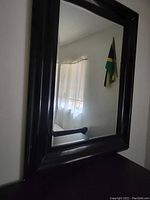 Black framed rectangular mirror hanging on a wall, showing partial reflection of a window with curtains and a flag.
