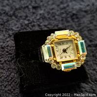 Close-up of a square-faced ring watch with rhinestones surrounding the dial and Roman numeral markers.