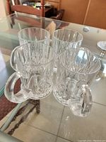 Four clear lead crystal coffee or tea mugs with intricate cut designs, handles and thick bases, placed on glass table.