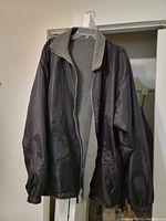Front view of men's fleece jacket showing outer nylon shell and inner fleece lining, hung on clear hanger against wall.
