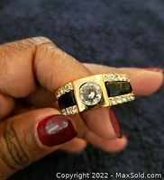 Close-up photo of a gold-tone men's ring held in hand, showing central clear cubic zirconia stone, black enamel inlay and rows of smaller clear CZ stones on sides