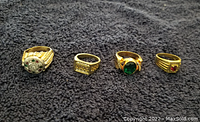 Five men's gold-tone rings laid out on a dark fabric background. First ring has cluster stones, second is a textured band, third has a large green stone, fourth has a small red stone.