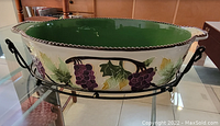 Photo of oval ceramic baking dish in black metal basket holder, showing grape vine decoration on outside and green interior.