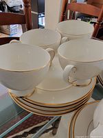 Overall view of four white ceramic tea cups stacked above four matching plates with gold rims on a glass table.