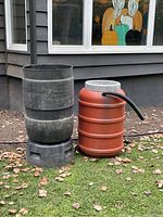 Two water barrels shown outdoors on grass; one black open barrel stacked on black stand, one red barrel with hose and faucet attachments