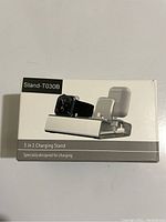 Photo of the product box showing a white charging stand with slots for three devices: smartwatch, smartphone, and earbuds. Box label reads Stand-T030B along with '3 in 1 Charging Stand Specially designed for charging'.