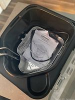 Photo showing the open fryer interior containing a perforated cooking basket with handle, a metal hook, and instruction manual folded inside.