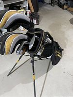 Image of the golf bag containing golf clubs with stand legs extended on concrete floor in storage area.
