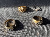 Four men's rings displayed on a textured dark fabric surface showing varied designs and stones.