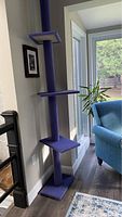 Full view of the tall purple carpeted cat tree, showing its placement near a wall and window, displaying three flat platforms and a square base.