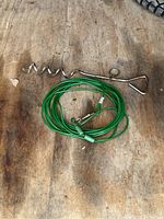 Photo shows a green-coated wire dog leash coiled with metal clip and connectors alongside a metal ground stake with helix screw design and triangular handle on a wooden surface.
