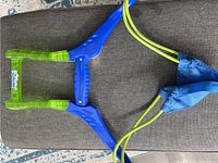 Full view of water balloon slingshot showing blue and green plastic frame with attached yellow elastic cords and blue slings.