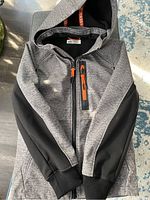 Front view of H&M youth spring coat in grey and black with hood and orange zip pulls
