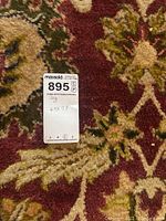 Close-up of area rug showing floral pattern and a slight worn edge with a MaxSold tag indicating approximate size
