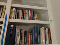 Two white shelves filled with novels and cookbooks, showing titles and spines clearly with some classic and contemporary books.