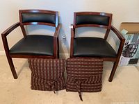 Two wood and black faux leather armchairs side by side, showing armrests and backrests with comfortable padding. Two striped cushions are placed on the floor in front of the chairs.