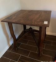 Front angle of square wooden table with ornate base showing wood grain top and carved legs on tile floor near corner wall.