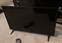 Front view of the Insignia 24 inch LED TV with one remote on the screen and other remote on the carpet in front.