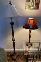 Photo showing both lamps side by side: the tall ornate metal floor lamp with beige shade and the shorter wood grain table lamp with dark brown shade on a glass table.