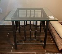 Side view of one end table showing textured metal base with decorative vertical rods and clear glass top.
