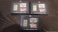 Three sealed boxed Coors Banquet pins sets on a dark surface showing front packaging and logos.