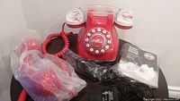 Phone shown with red body, white rotary-style push buttons, Coca-Cola logo in center, one handset wrapped in plastic, manual, and original packaging visible.