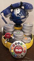 Full view of the M&M's themed studio phone with the globe handset holder, yellow film reel base, silver film canisters, rotary dial, and red M&M figurine.