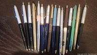 Overview photo showing 16 vintage promotional pens of various colors and conditions laid out in a row to illustrate the full lot content.