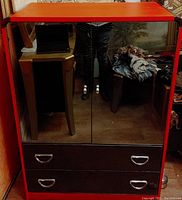 Front view of red and black dresser with mirrored double glass doors and two drawers below, showing handles and shiny surfaces reflecting surroundings.