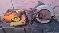Lot of three power tools displayed on a black case, including a Skilsaw circular saw, a yellow cordless drill, and an orange corded drill.