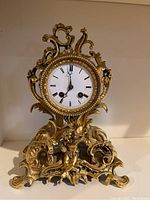 Front view of the gold-tone ornate metal mantel clock showing detailed scroll, floral, and cherub design with white clock face and Roman numerals.