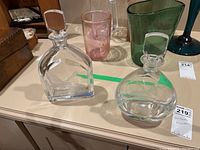 Two clear glass Orrefors decanters, approximately 10-11 inches tall, sit on a table with a variety of other items in the background.