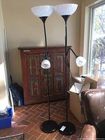 Full view of two black standing floor lamps with white bowl-shaped upper lamps and adjustable smaller lower lamps in front of wooden cabinet and window.