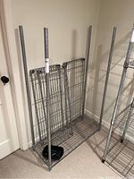 Disassembled metal bakers rack shelf unit showing wire grid shelving and tubular metal posts against a beige wall.