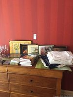Overview photo showing stacks of placemats, folded linen napkins, and paper napkins arranged on a wooden dresser with a red striped wall in background.