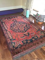 Rectangular rug laid on hardwood floor showing red/orange field with central medallion and navy border, tassel-fringed ends visible