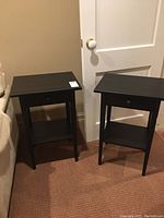 Pair of black wooden side tables shown next to a door and couch, featuring one mounted drawer and open lower shelf.