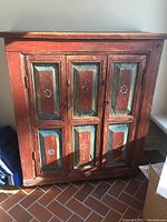 Front view of vintage wooden cabinet showing distressed red and teal painted panels with floral motifs.