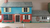 Front exterior view of the Marx tin metal dollhouse showing the two-story house with detailed brick and siding paint, windows with shutters, door, and attached garage with floral painted side.