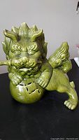 Green glazed ceramic Asian dog figurine with dagger in mouth, viewed from front left showing detailed facial features and spherical base with cutouts.