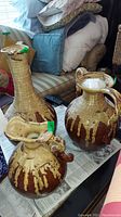 Three distressed ceramic jugs arranged on a newspaper-covered table, showing varying shapes and sizes with pale yellow and brown finishes and visible wear.