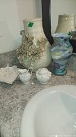 Overview of assortment including floral ceramic vase with flowers, small marbled vase and white ceramic decorative pieces on bathroom counter near sink.