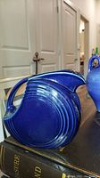 Large blue ceramic pitcher with circular ridged detail and handle, displayed on books.