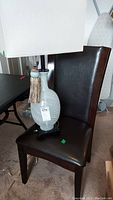 Glass and metal lamp on a dark vinyl and wood chair. The lamp has a light blue-gray ribbed glass base and rectangular cream-colored fabric shade with tassel.