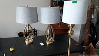 Photo of two matching metal and glass table lamps with light gray fabric shades, and a floor lamp with white shade on a black surface with surrounding furniture.